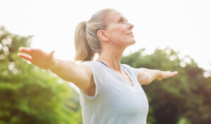 Lächelnde Frau im Freien mit ausgestreckten Armen, trägt ein weißes Hemd, im Hintergrund Grün.