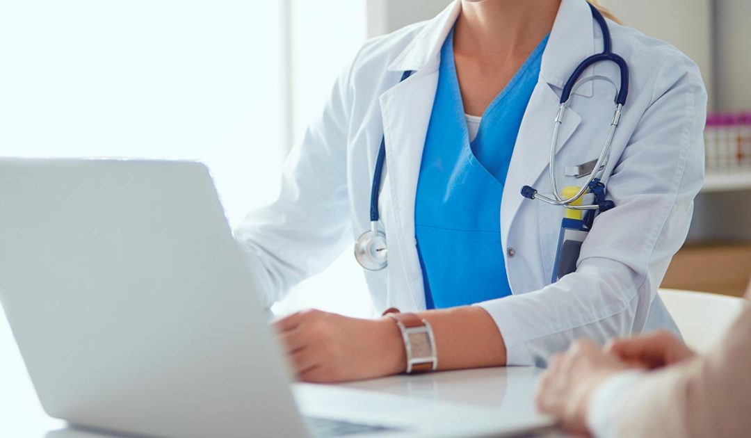 Ein Arzt im weißen Kittel mit Stethoskop und Laptop spricht mit einem Patienten über den Schreibtisch hinweg.