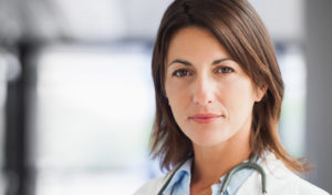 Eine selbstbewusste Ärztin mit braunem Haar und Stethoskop, die direkt in die Kamera blickt.