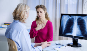 Eine Frau berührt ihre Brust, während sie mit einem Arzt spricht, während auf einem Computerbildschirm eine Röntgenaufnahme der Brust angezeigt wird.