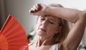 Ältere Frau mit geschlossenen Augen, die sich Luft zufächelt und ihre Stirn abwischt, sie wirkt heiß oder überhitzt.