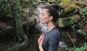 Frau steht im Freien mit geschlossenen Augen, Hände auf der Brust, vor einem Wasserfall und viel Grün.