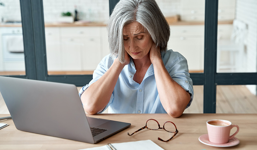 Eine ältere Frau sitzt mit einem Laptop an einem Tisch, sieht gestresst aus und hält ihren Hals, eine Brille und Kaffee in der Nähe.