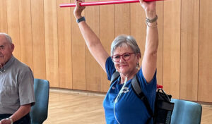 Lächelnde ältere Frau hebt im Innenbereich eine rote Gymnastikstange über den Kopf; andere sitzen in der Nähe.