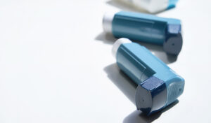 Zwei blaue Asthma-Inhalatoren liegen auf einer weißen Oberfläche mit Licht und Schatten.