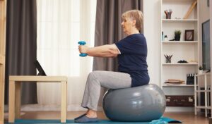 Eine ältere Frau sitzt auf einem Gymnastikball, stemmt Hanteln und schaut dabei in einem gemütlichen Wohnzimmer auf ein Tablet.