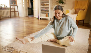 Ältere Frau sitzt in gemütlicher Kleidung auf einem Teppich im Wohnzimmer und macht Dehnübungen vor einem Laptop, während eines Online-Sportkurses.