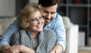 Eine lächelnde ältere Frau mit Brille wird von einem jüngeren Mann umarmt; beide sitzen zusammen zu Hause auf einem Sofa.