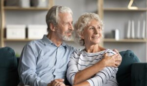 Lächelndes älteres Paar sitzt auf einem Sofa, umarmt sich und blickt glücklich in die Ferne.