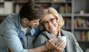 Lächelnde ältere Frau mit einer Tasse, in eine Decke gehüllt, von einem jüngeren Mann in einem gemütlichen Raum umarmt.