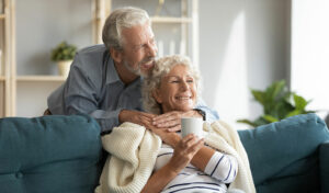 Lächelndes älteres Paar sitzt auf einem Sofa, einer hält eine Tasse, und genießt einen gemütlichen Moment zusammen zu Hause.