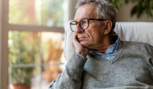 Ein älterer Mann mit Brille blickt nachdenklich aus dem Fenster und stützt sein Kinn auf seine Hand.