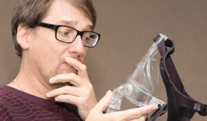 Mann mit Brille betrachtet nachdenklich eine CPAP-Maske in seiner Hand vor einem schlichten Hintergrund.