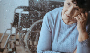Traurig blickende ältere Frau am Fenster, im Hintergrund ein Rollstuhl.