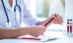 Ein Arzt untersucht in einem Labor ein Fläschchen mit Blut. Auf dem Tisch liegen ein Klemmbrett und Blutproben.