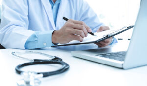 Arzt schreibt auf einem Klemmbrett an einem Schreibtisch mit einem Stethoskop und einem Laptop in der Nähe.