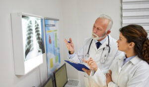 Zwei Ärzte besprechen eine Röntgenaufnahme der Brust, die auf einem Leuchtkasten in einer Arztpraxis angezeigt wird.