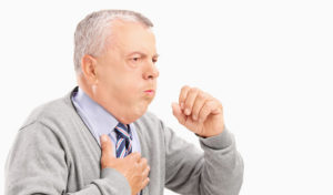 Älterer Mann in einem grauen Pullover, der hustet und seine Brust hält, er scheint sich unwohl zu fühlen.