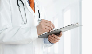 Arzt im weißen Kittel hält ein Klemmbrett in der Hand und schreibt, mit einem Stethoskop um den Hals.