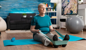 Ältere Frau trainiert zu Hause auf einer Yogamatte und verwendet ein Widerstandsband um ihre Füße.