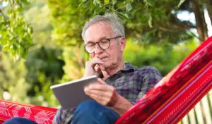 Ein älterer Mann mit Brille sitzt in einer Hängematte und hält ein Tablet in der Hand