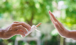 Eine Person bietet eine Zigarette an, während eine andere mit einer Handbewegung ablehnt; im Hintergrund ist Grün zu sehen.