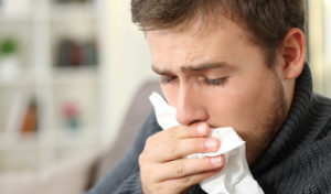 Ein Mann im Pullover hält sich ein Taschentuch vor die Nase und sieht aus, als würde er niesen oder erkältet sein.