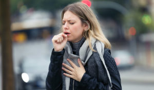 Frau im Freien hustet in ihre Faust, mit einer Hand auf der Brust, trägt einen Schal und eine Jacke.