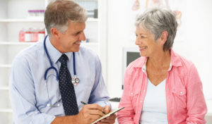 Männlicher Arzt spricht in einer Arztpraxis mit einer älteren Patientin und lächelt sie an.