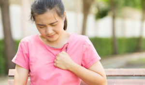 Frau in einem rosa Hemd sitzt auf einer Bank, hält sich die Brust und scheint Schmerzen oder Unbehagen zu haben.