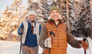 Lächelndes älteres Paar beim Langlauf im verschneiten Wald, warm gekleidet in Winterkleidung und Mützen.