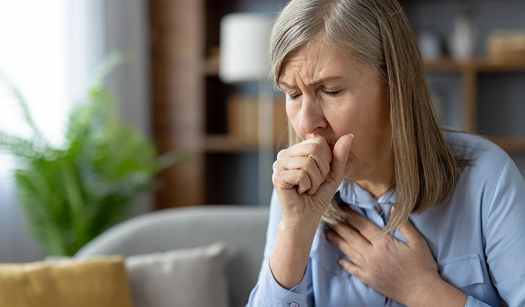 Ältere Frau, auf Sofa sitzend, hat einen Hustenanfall, und hält sich die Hand vor den Mund