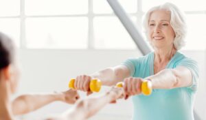 Lächelnde ältere Frau in einem hellblauen Hemd, die mit einem Partner in einem hellen Raum gelbe Hanteln hebt.