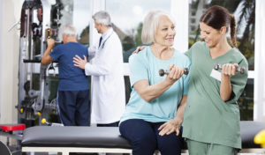 Ältere Frau hebt mit Hilfe eines Pflegers eine Hantel; im Hintergrund trainieren zwei Männer.