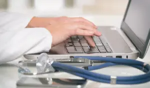 Hände tippen auf einer Laptoptastatur, während daneben ein Stethoskop auf dem Schreibtisch liegt.