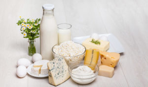 Verschiedene Milchprodukte, darunter Milch, Käse, Eier, Butter und Joghurt, angeordnet auf einer hellen Holzoberfläche.