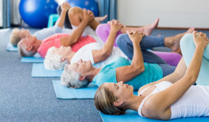 Eine Gruppe von Senioren und eine jüngere Frau liegen auf Yogamatten und strecken sich mit einem an die Brust gezogenen Knie.