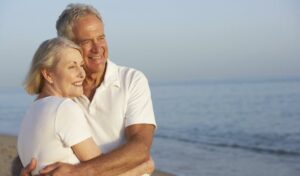 Lächelndes älteres Paar in weißer Kleidung umarmt sich an einem Strand mit dem Meer im Hintergrund.