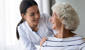 Eine junge Pflegekraft lächelt eine ältere Frau an und stützt sie, die dasitzt und zurücklächelt.