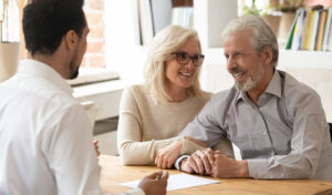 Lächelndes älteres Paar hält Händchen und unterhält sich mit einem Fachmann an einem Schreibtisch in einem hellen Büro.