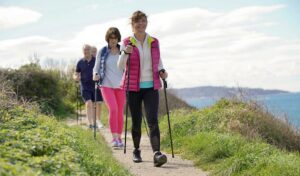 Drei Personen wandern mit Trekkingstöcken auf einem Küstenpfad mit viel Grün und dem Meer im Hintergrund.
