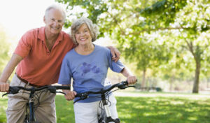 Lächelndes älteres Paar mit Fahrrädern steht in einem sonnigen Park mit grünen Bäumen im Hintergrund.