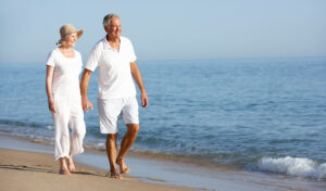Ein älteres Paar in weißer Kleidung geht Hand in Hand an einem sonnigen Strand in der Nähe des Wassers entlang.