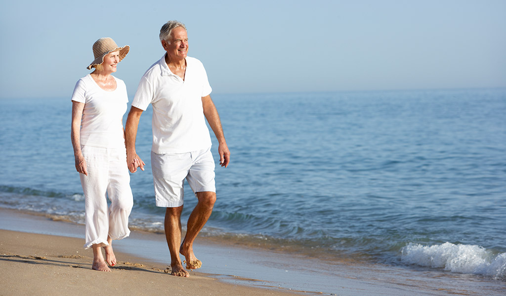 Ein älteres Paar in weißer Kleidung geht Hand in Hand an einem sonnigen Strand in der Nähe des Wassers entlang.