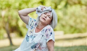 Ältere Frau im Freien wischt sich an einem sonnigen Tag mit einem Tuch den Schweiß ab und sieht überhitzt und müde aus.