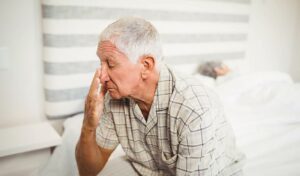 Ein älterer Mann sitzt auf dem Bett und reibt sich die Augen, im Hintergrund schläft eine andere Person.