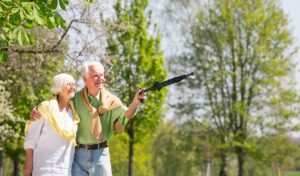 Lächelndes älteres Paar geht Arm in Arm im Freien spazieren; ein Mann zeigt mit geschlossenem Regenschirm an einem sonnigen Tag.