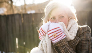 Lächelnde ältere Frau in Winterkleidung hält an einem sonnigen Tag draußen einen Schal vors Gesicht.