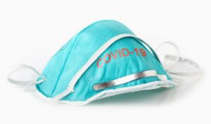 Eine blaue Gesichtsmaske mit der Aufschrift „COVID-19“ mit weißen Bändern und einem metallischen Nasenstreifen auf weißem Hintergrund.