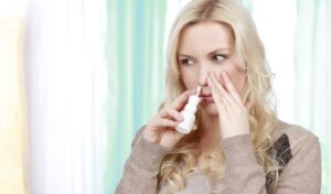 Eine Frau benutzt ein Nasenspray, während sie ihre Nase zuhält, und steht mit nachdenklichem Gesichtsausdruck im Haus.